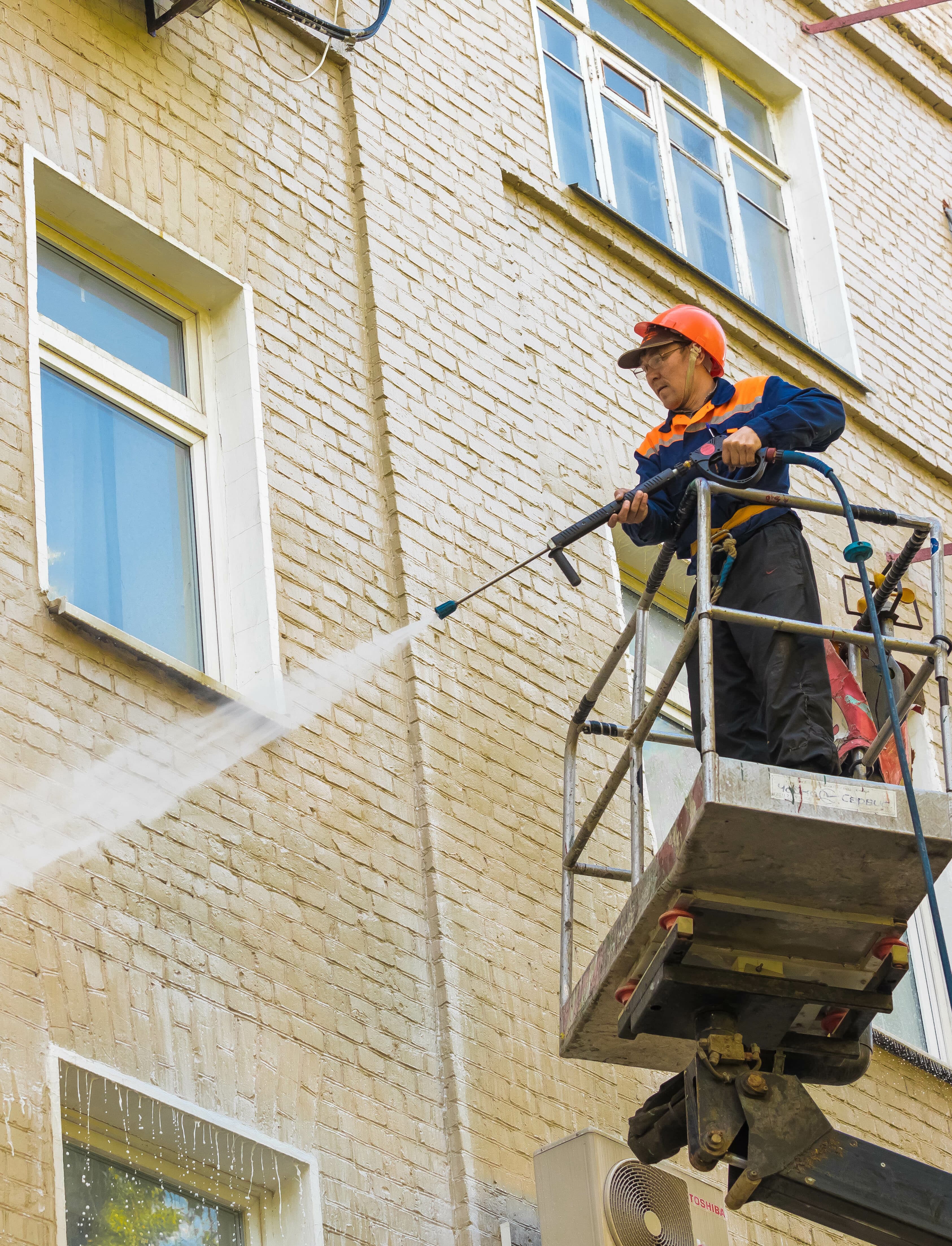 The employee of municipal services washes a facade of the multi-storey building by means of the high-pressure apparatus. Brick Cleaning