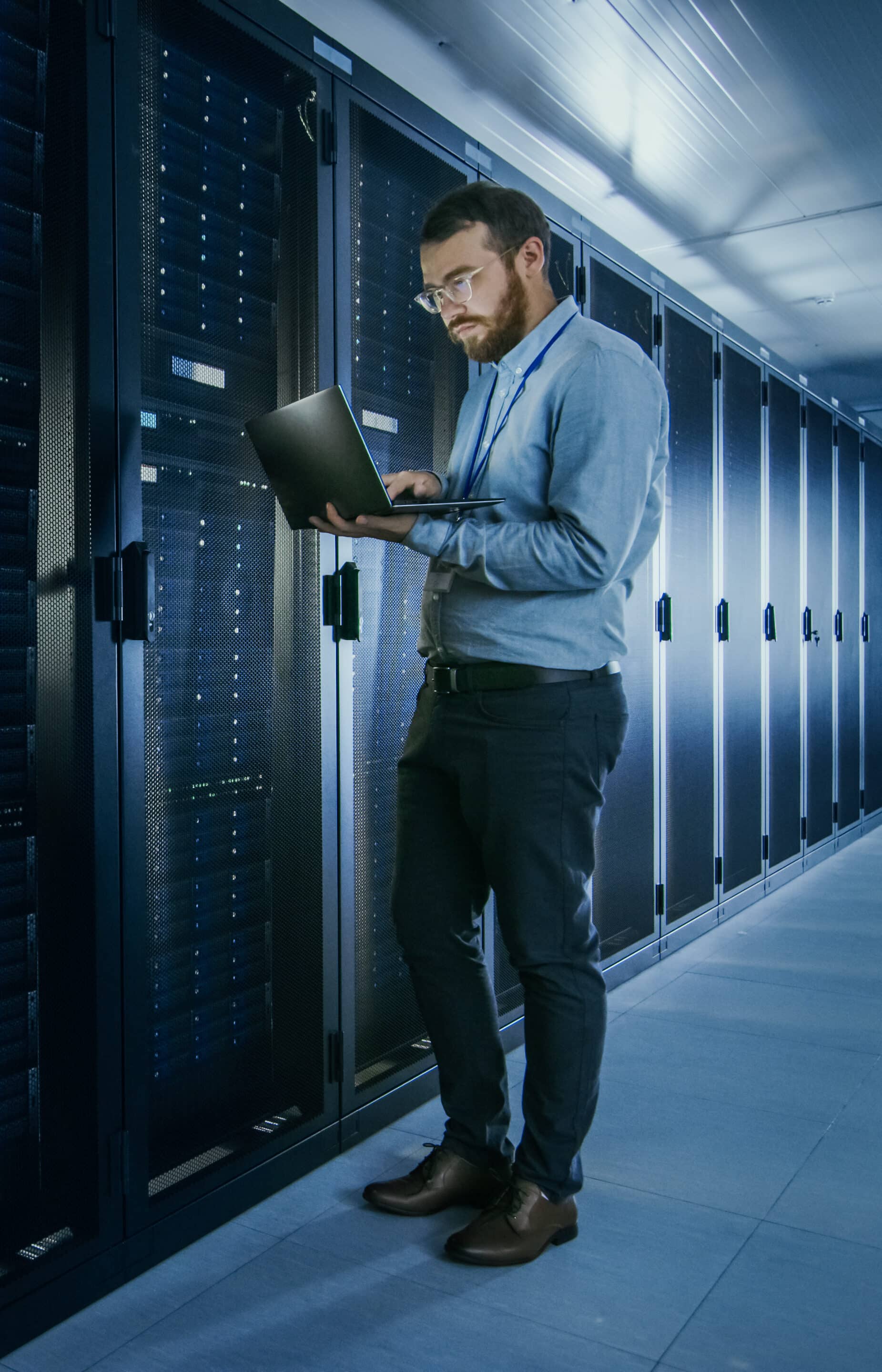 Bearded Specialist VOIP Systems in Glasses is Working on Laptop in Data Center while Standing Before Server Rack. Running Diagnostics or Doing Maintenance Work.