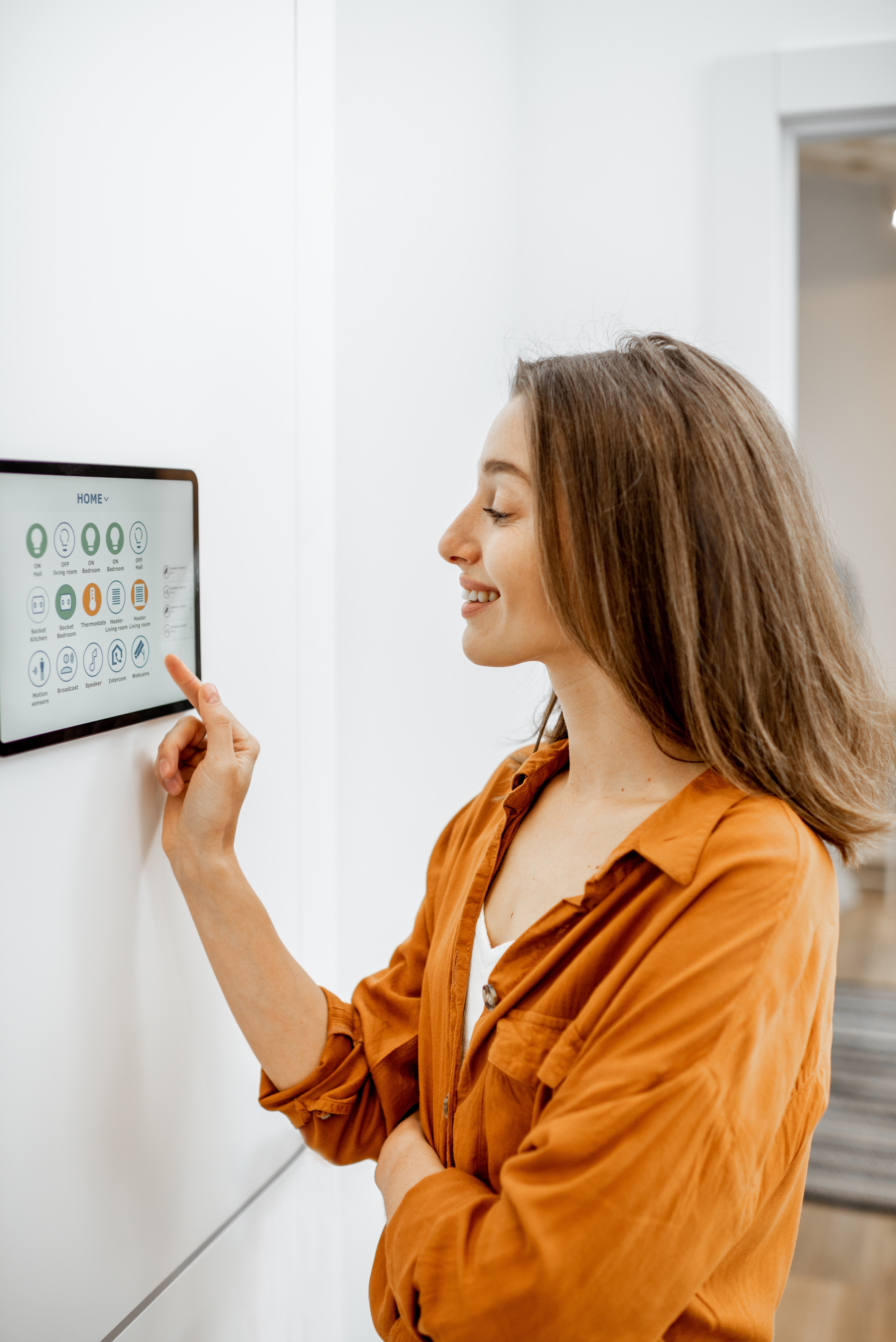 Young woman controlling home with a digital touch screen panel installed on the wall in the living room. Concept of a smart home and mobile application for managing smart devices at home