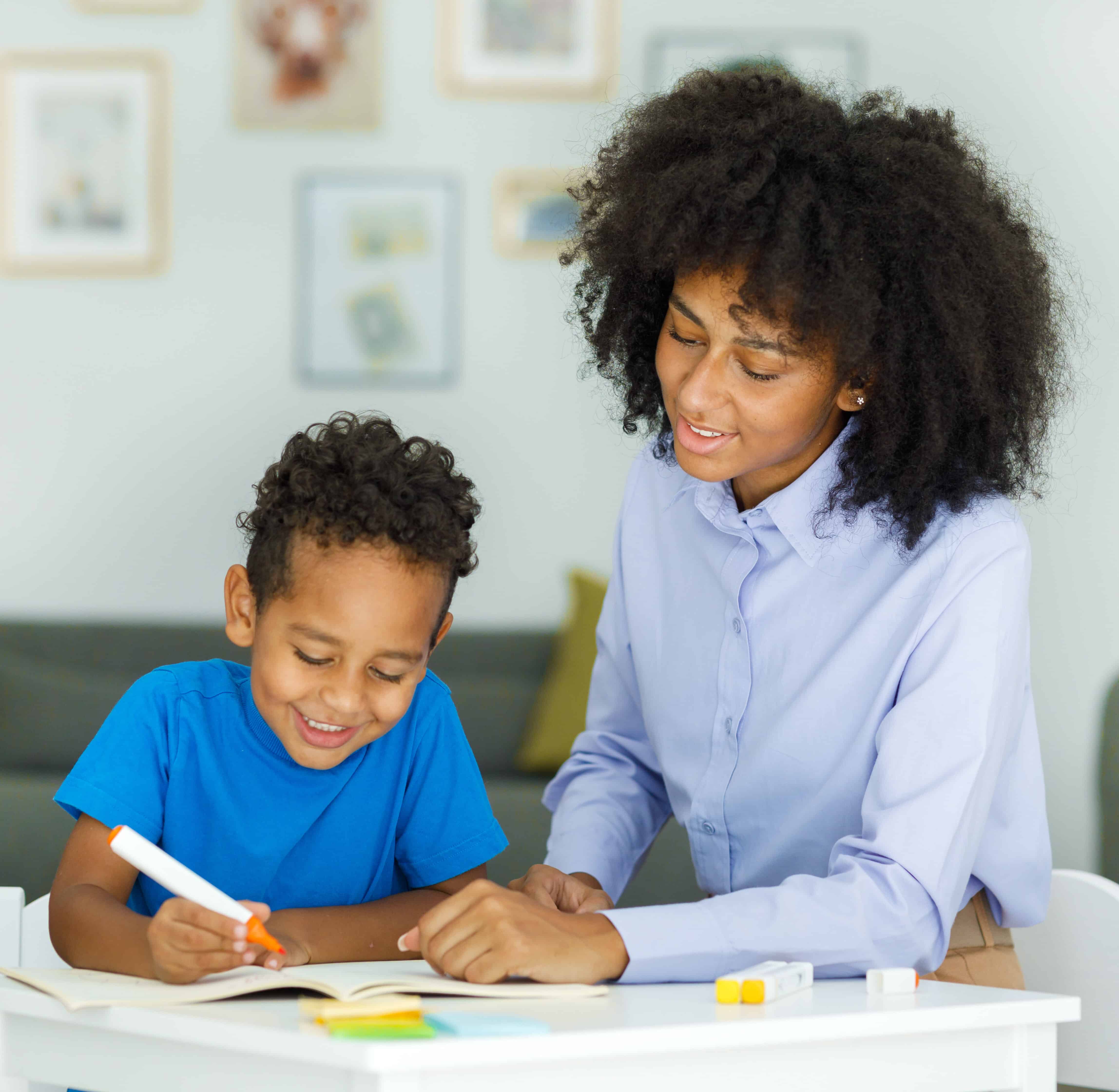 Female infant school Spanish Tutor working one on one with a young schoolboy, sitting at a table writing in a classroom, front view. High quality photo