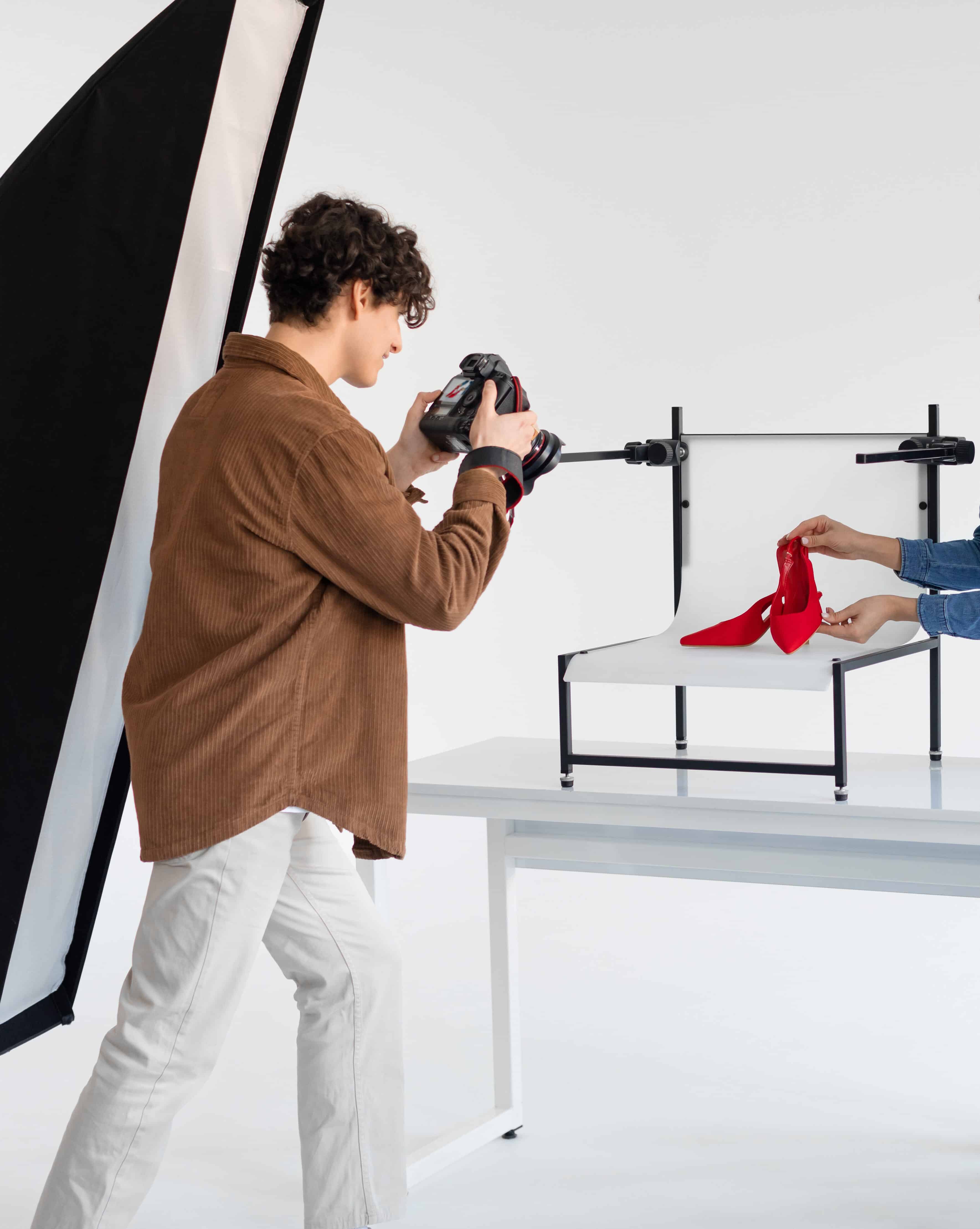 Young male photographer taking photo of stylish red shoes on white platform background in photostudio with modern equipment, lady helping to make content photoshoot