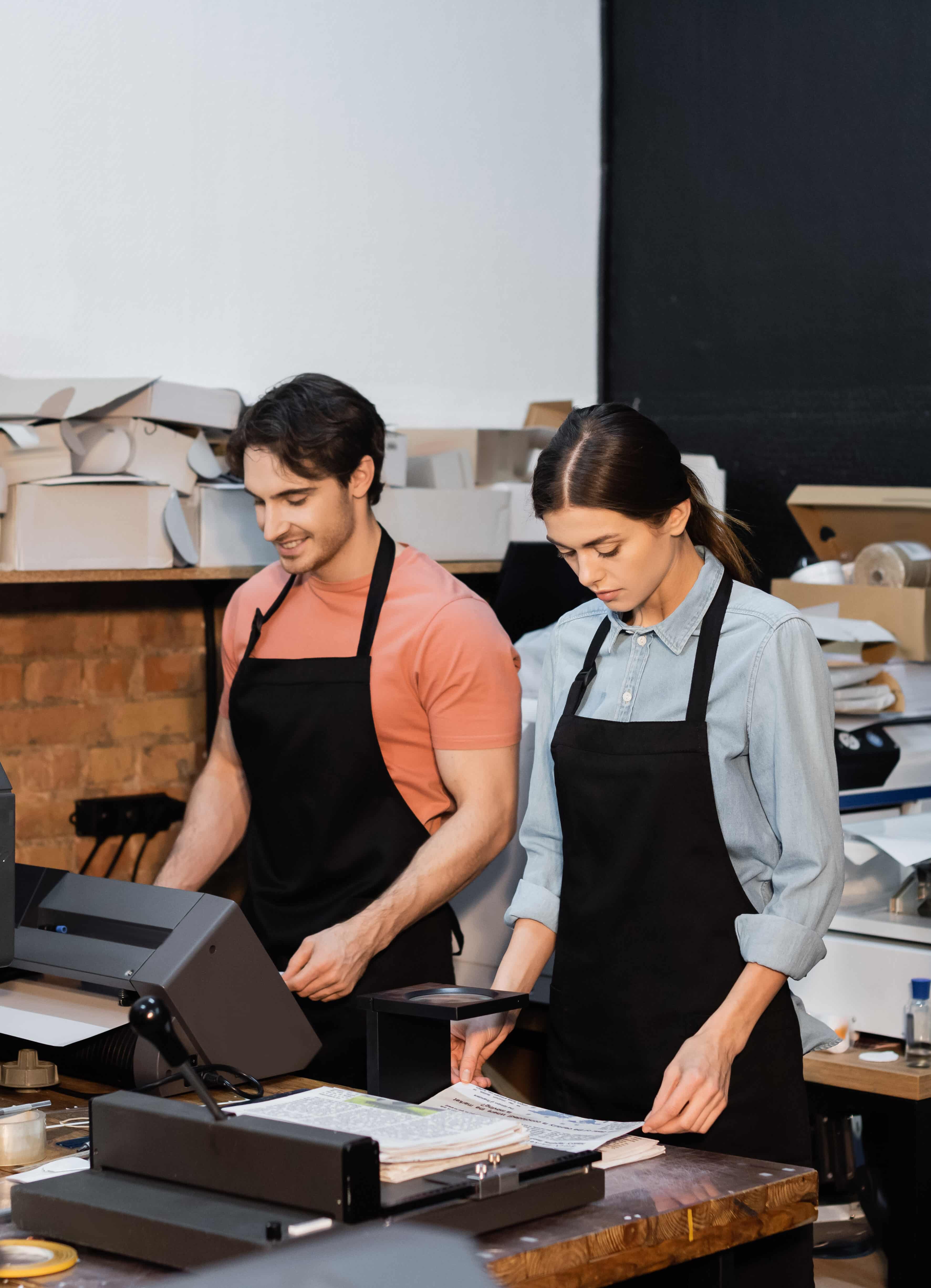 Cheerful man in apron working with printer next to colleague in print