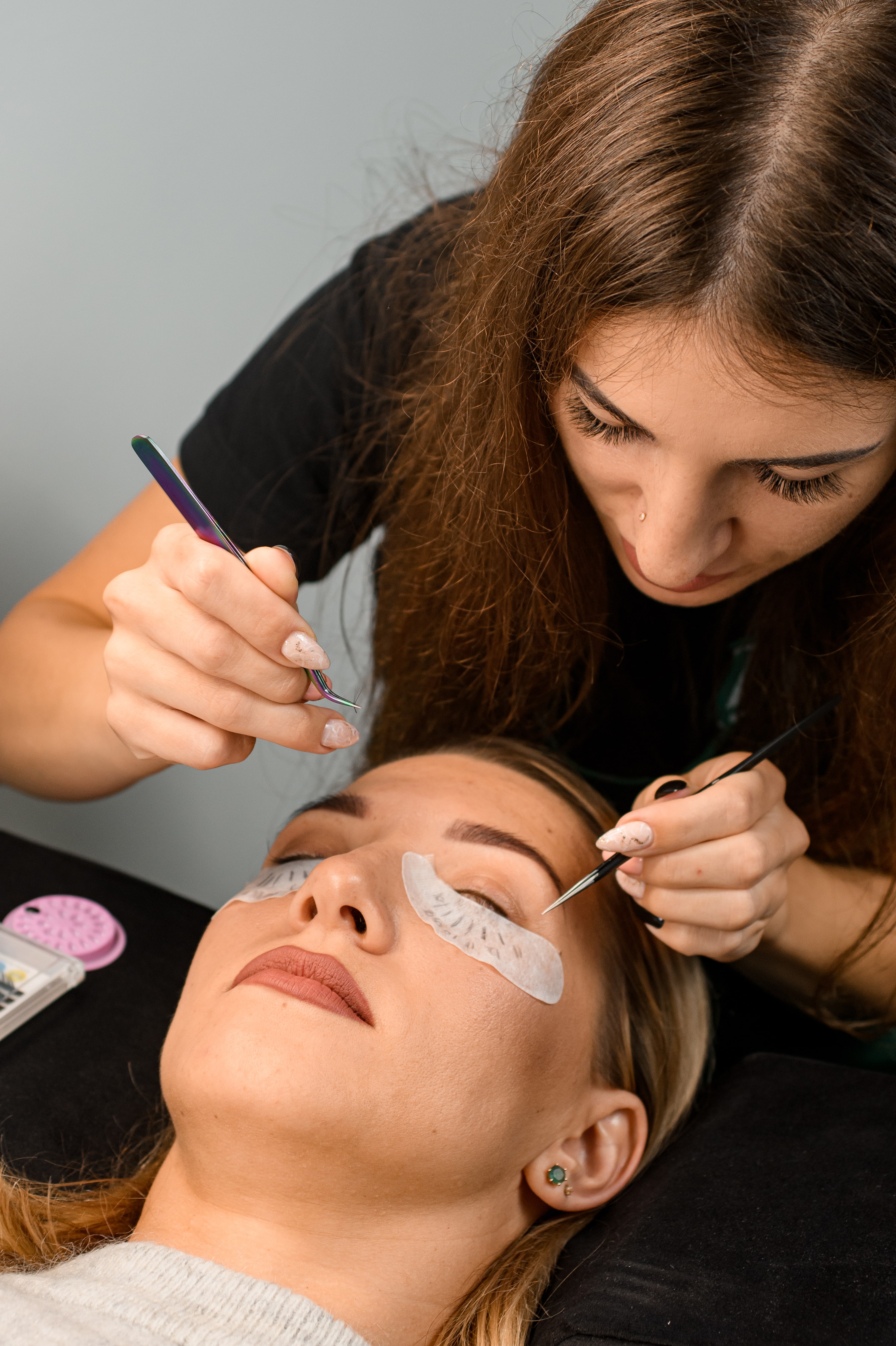 Female cosmetologist with tweezers attaches artificial eyelashes extensions to female client at salon.