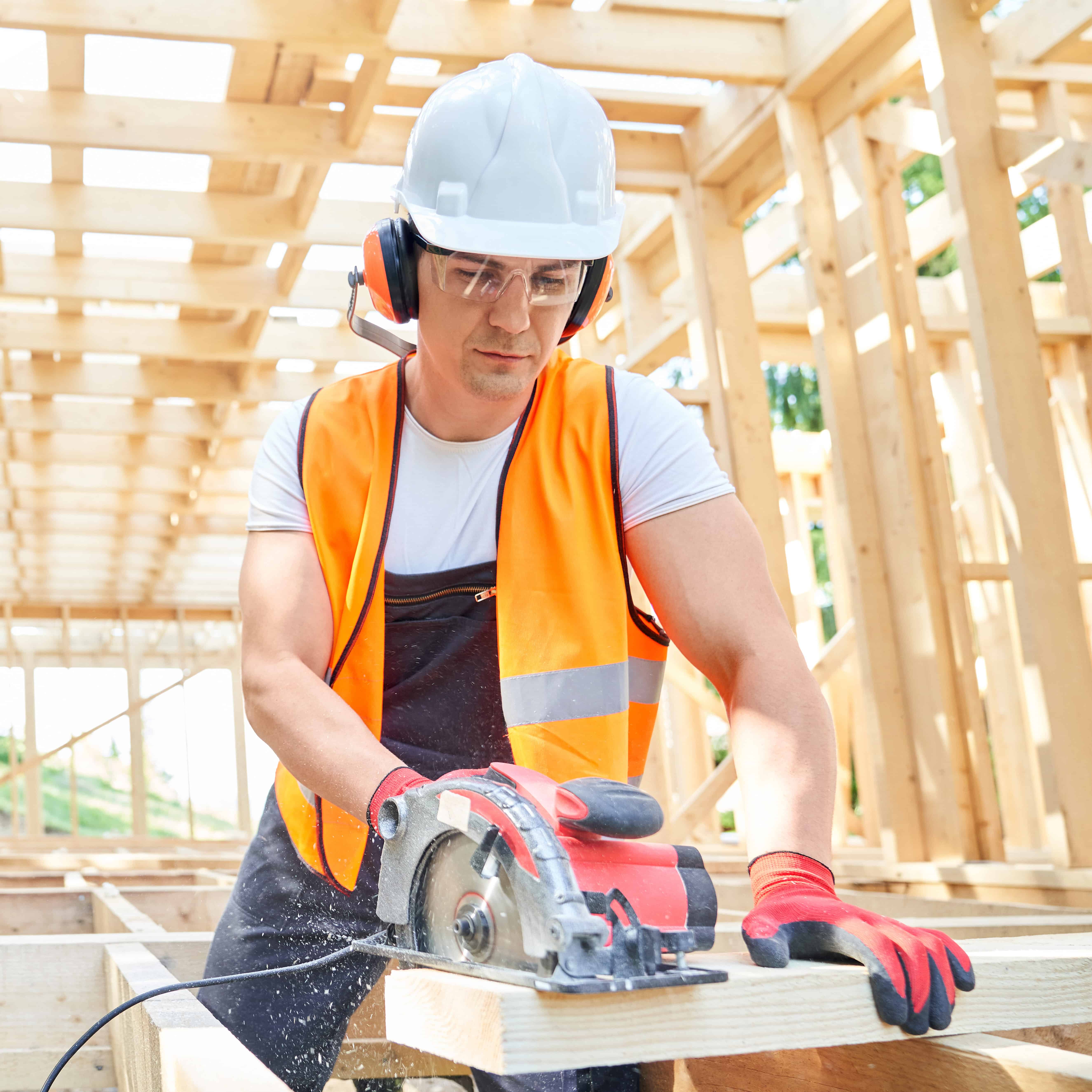 Front view of craftsman, builder standing, looking down, cutting wodden gilder by professional equipment. Builder wearing yellow vest, helmet, glasses working. Concept of a Timber Roofing