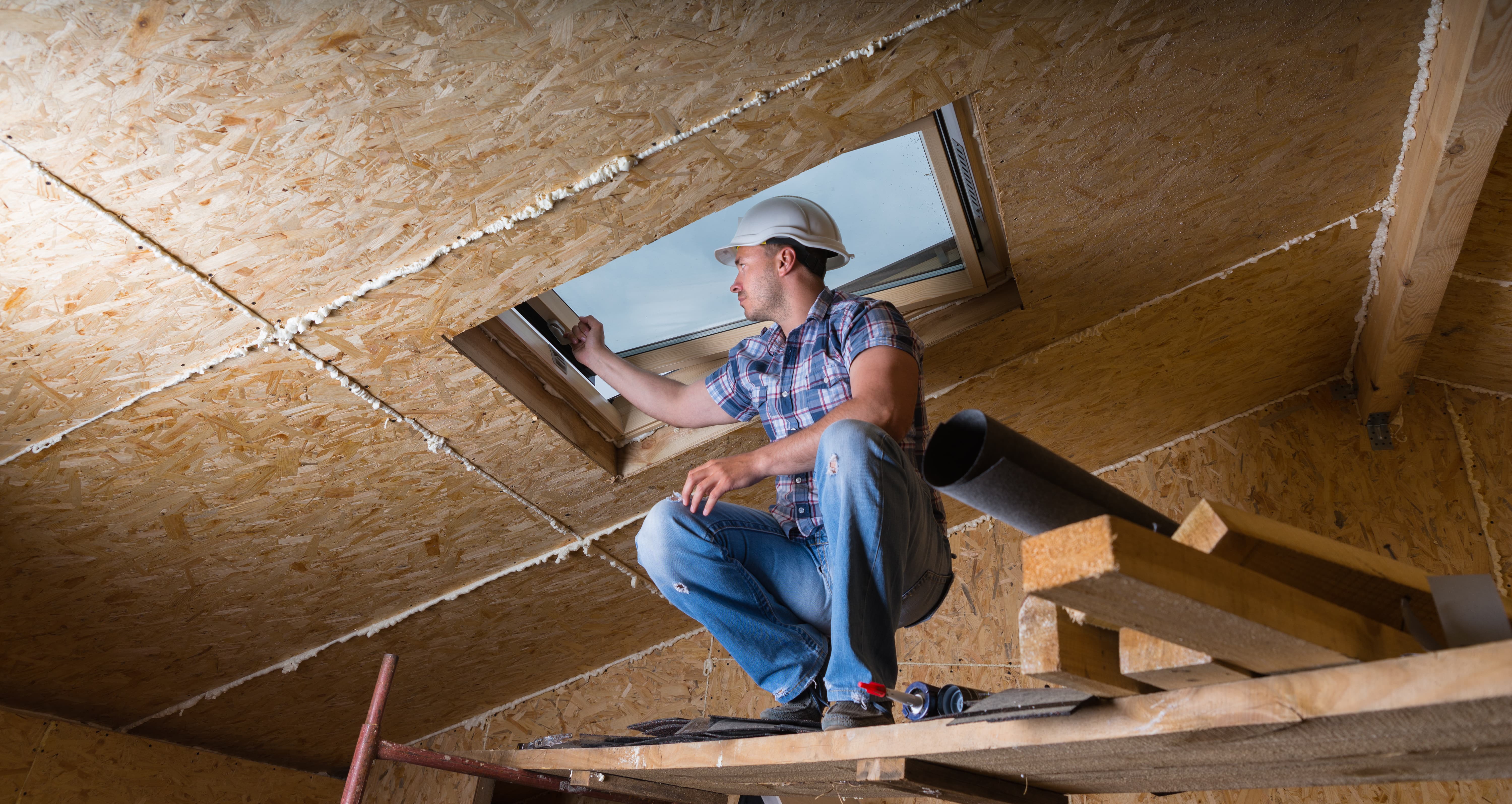 Low Angle View of Male Construction Worker Builder Crouching on Elevated Scaffolding near Ceiling and Inspecting Frame of Sky Light Window in Unfinished House with Exposed Particle Plywood Board