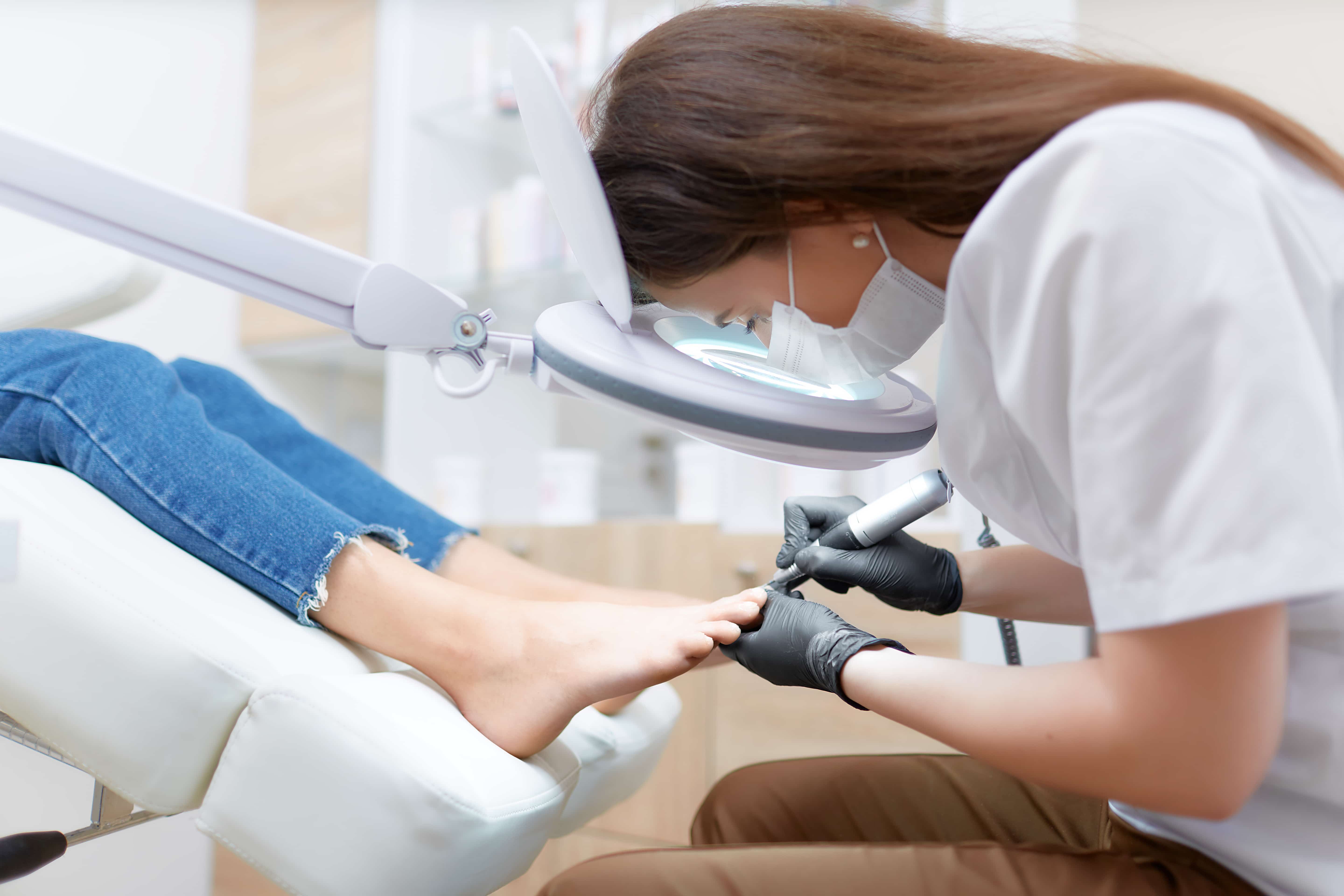 View of Chiropodist doctor in white working at beauty salon, making polish procedure for foot with special equipment for blondie client. Young woman resting, caring about her health and foot.