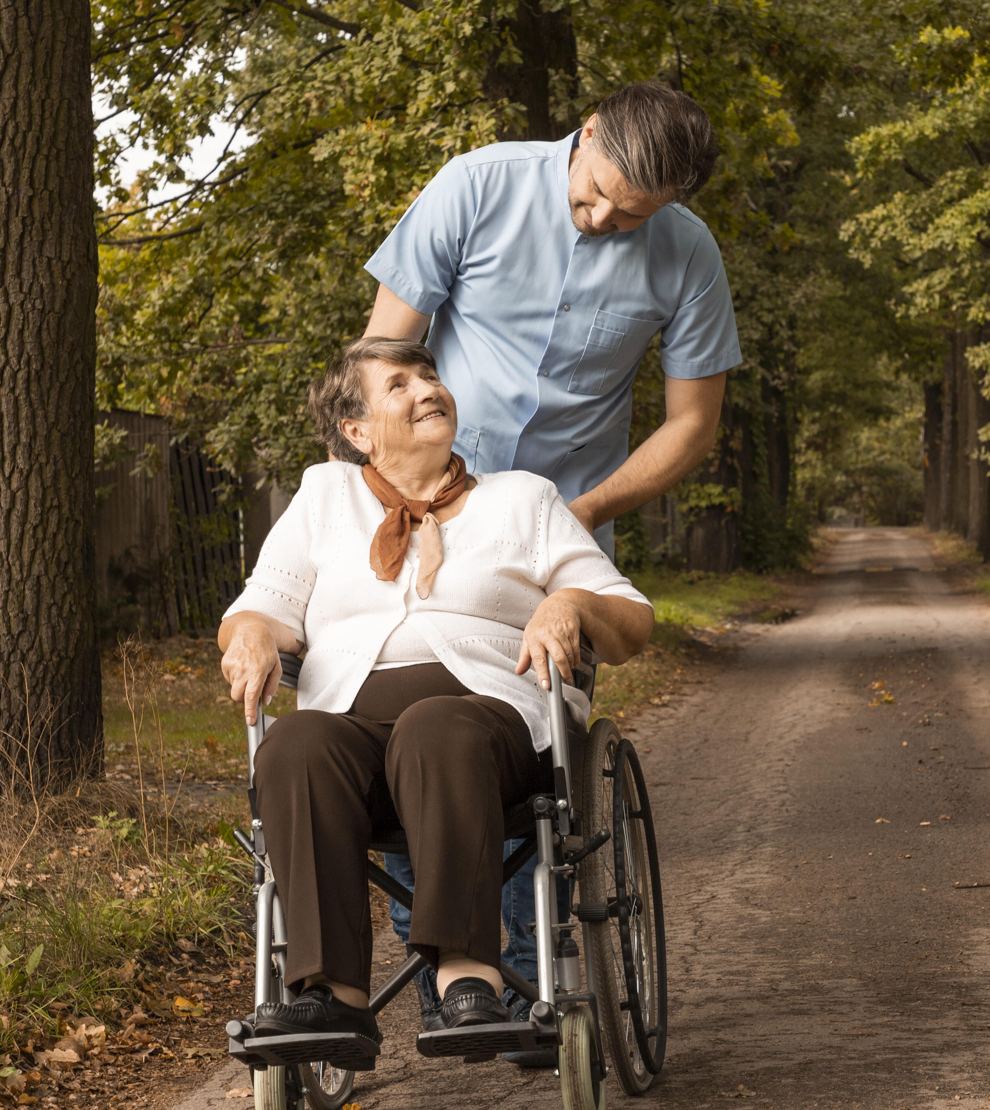 Male nurse taking for a stroll in park female senior in a wheelchair doing some Disability Services