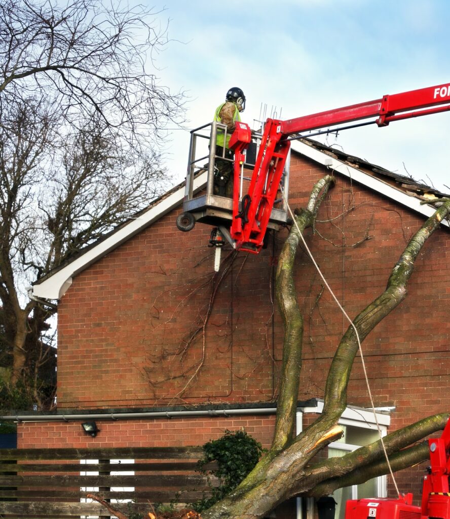 Tree surgeon working up cherry picker repairing storm damaged roof after an uprooted tree fell on top of a residential house