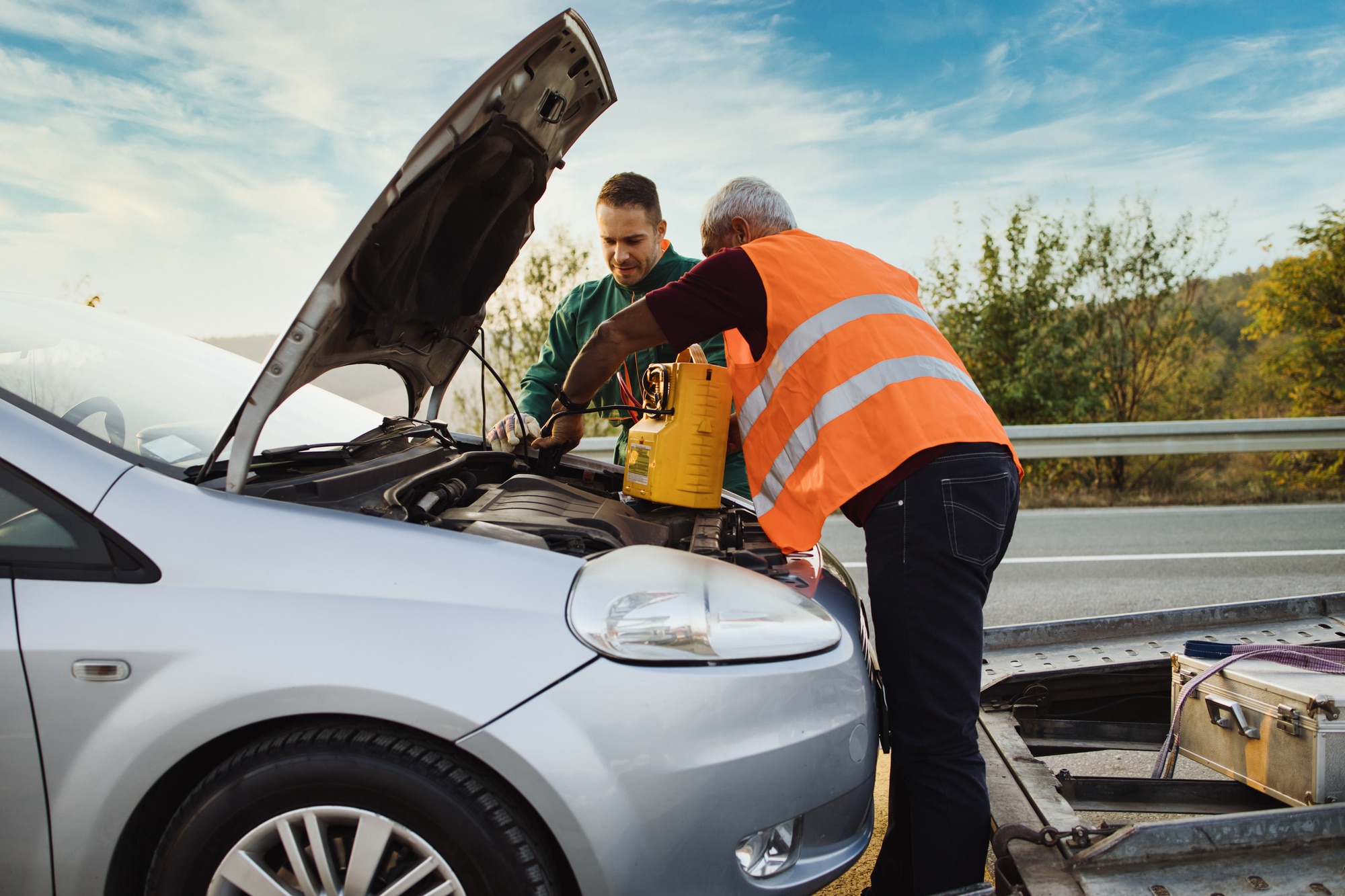 Two road assistant workers in towing service trying to start car engine with jump starter and energy station with air compressor. Roadside assistance concept.