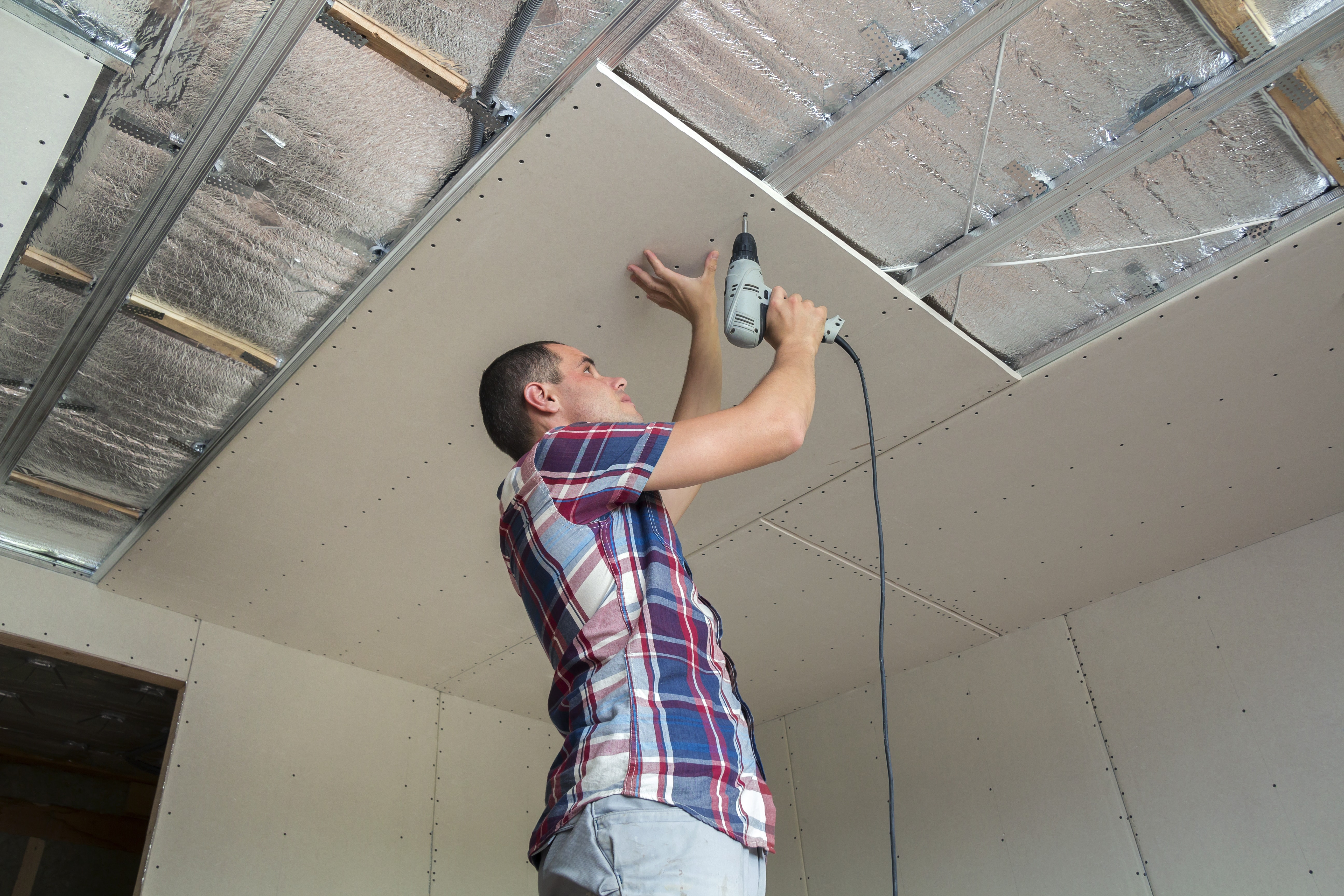 Young man in usual clothing and work gloves fixing drywall suspended ceiling to metal frame using electrical screwdriver on ceiling insulated with shiny aluminum foil.